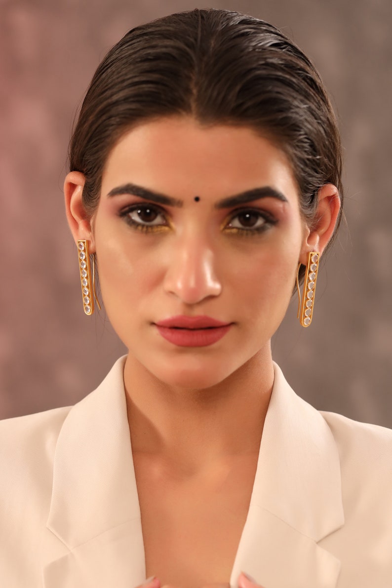 May include: A woman wearing a white blazer with gold earrings that have clear stones. The earrings are long and rectangular.