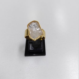 May include: A gold ring with a clear quartz stone set in the center. The stone is irregularly shaped and has a rough texture.