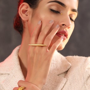 May include: A woman's hand wearing two gold rings. The ring on the index finger is a thin, long, pointed design. The ring on the thumb is a chunky, textured design.