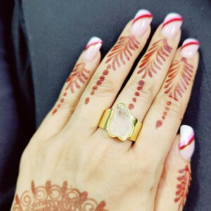 May include: A gold ring with a clear, irregular shaped stone set in the center. The ring is on a hand with henna designs and white and red nail polish.