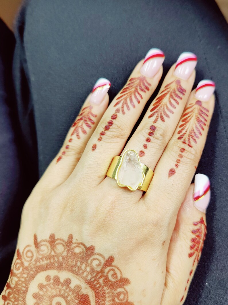 May include: A gold ring with a clear, irregular shaped stone set in the center. The ring is on a hand with henna designs and white and red nail polish.