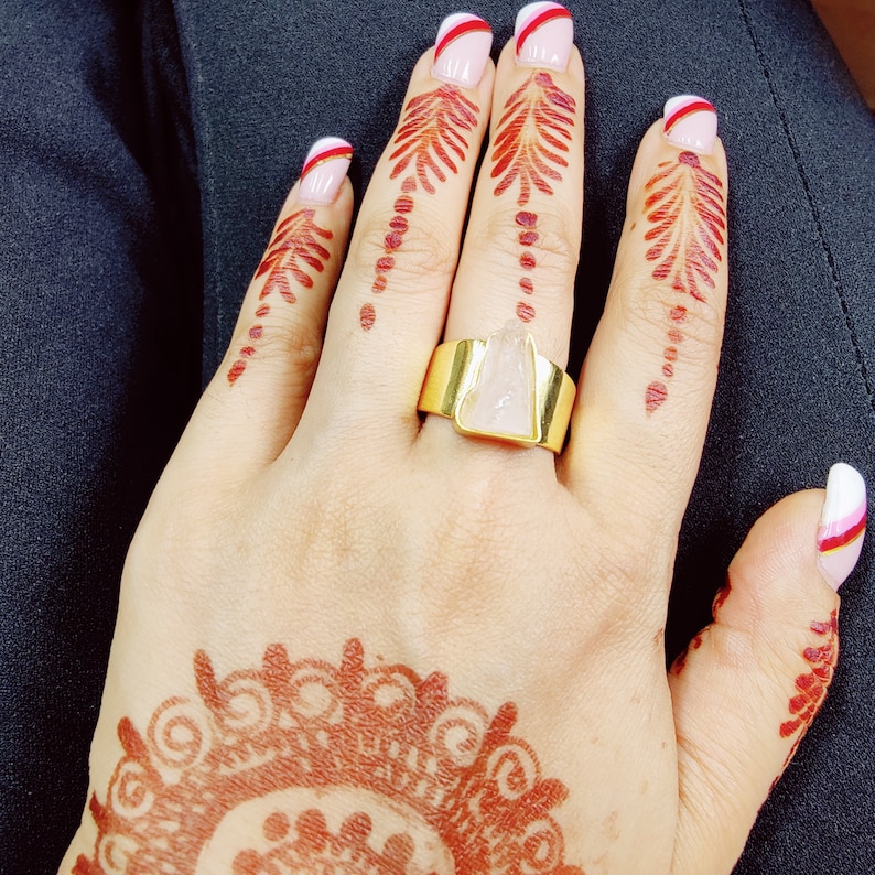 May include: A hand with henna designs and pink and white nail polish is wearing a gold ring with a clear, triangular stone.