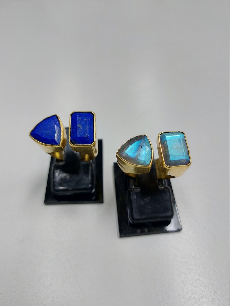Pu&ograve; includere: Due paia di anelli d'oro con pietre preziose blu e verdi. Gli anelli sono esposti su supporti neri.