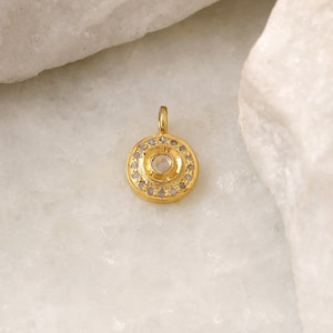 May include: A gold pendant with a round, clear gemstone surrounded by smaller gemstones. The pendant is on a white background.