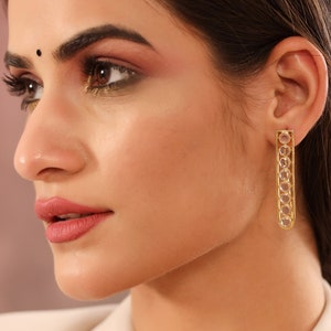 May include: Gold earrings with a long, rectangular design. The earrings are adorned with clear, round stones set in a gold frame.