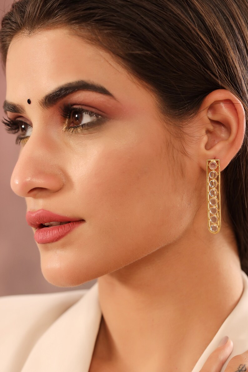 May include: Gold earrings with a long, rectangular design. The earrings are adorned with clear, round stones set in a gold frame.