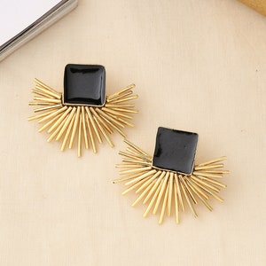 May include: A pair of gold-colored earrings with a black square center and a sunburst design. The earrings have a post back and are set against a neutral background.