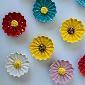 Puede incluir: Diez platos de cerámica con forma de margarita en varios colores, incluyendo rosa, blanco, azul verdoso, amarillo y rojo. Cada plato tiene un centro amarillo con un detalle de botón marrón.