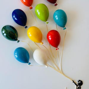 May include: A collection of eleven glossy, colorful balloons in shades of orange, red, green, blue, yellow, and white. The balloons are held by a silhouette of a child, creating a whimsical and artistic display.