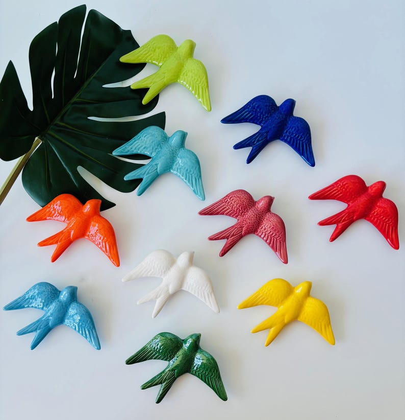 May include: A collection of ceramic swallow figurines in various colors, including green, blue, red, orange, white, and yellow. The birds are arranged on a white surface with a large green leaf in the background. Each bird has detailed wing and tail features.