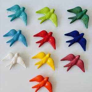 May include: Nine ceramic swallows in various colors, including teal, green, blue, red, white, yellow, burgundy, and orange. Each bird is depicted in flight, with outstretched wings, and is arranged on a white surface.