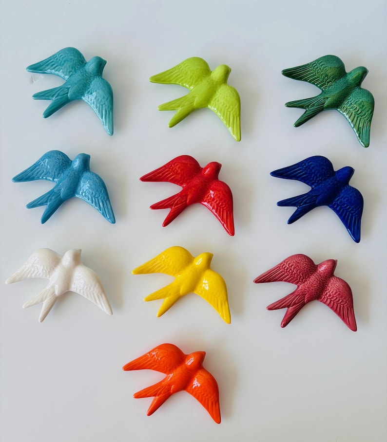 May include: Nine ceramic swallows in various colors, including teal, green, blue, red, white, yellow, burgundy, and orange. Each bird is depicted in flight, with outstretched wings, and is arranged on a white surface.