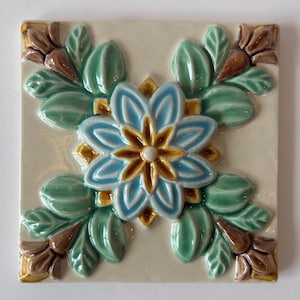 May include: Square ceramic tile with a raised floral design. The central flower is light blue with a yellow center, surrounded by green leaves and brown accents. The tile has a cream-colored background.