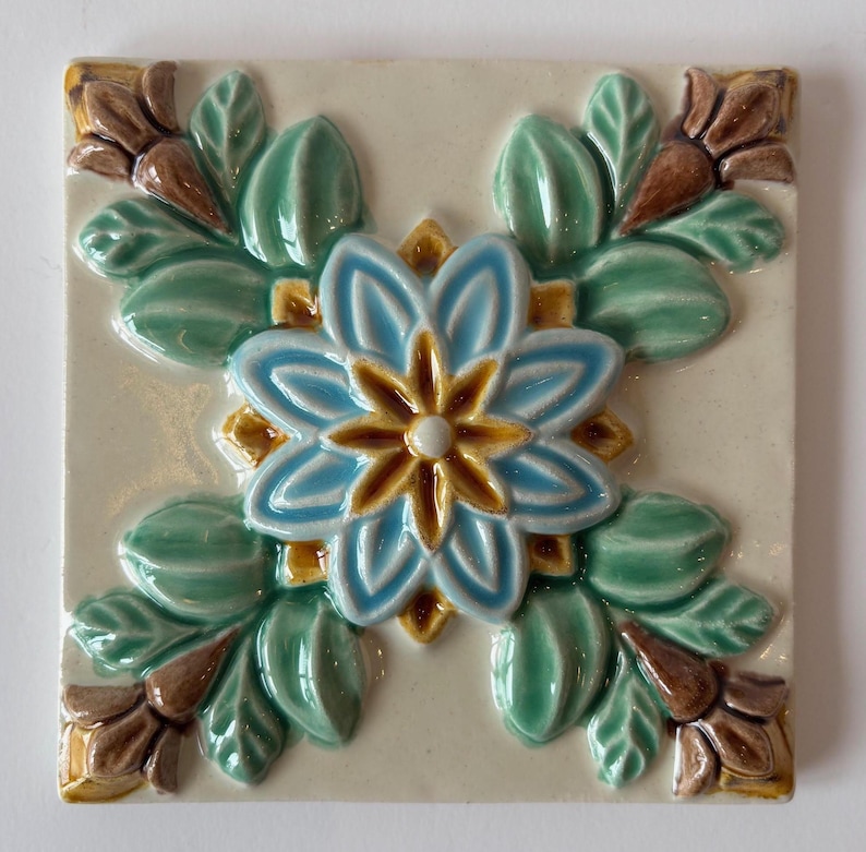 May include: Square ceramic tile with a raised floral design. The central flower is light blue with a yellow center, surrounded by green leaves and brown accents. The tile has a cream-colored background.
