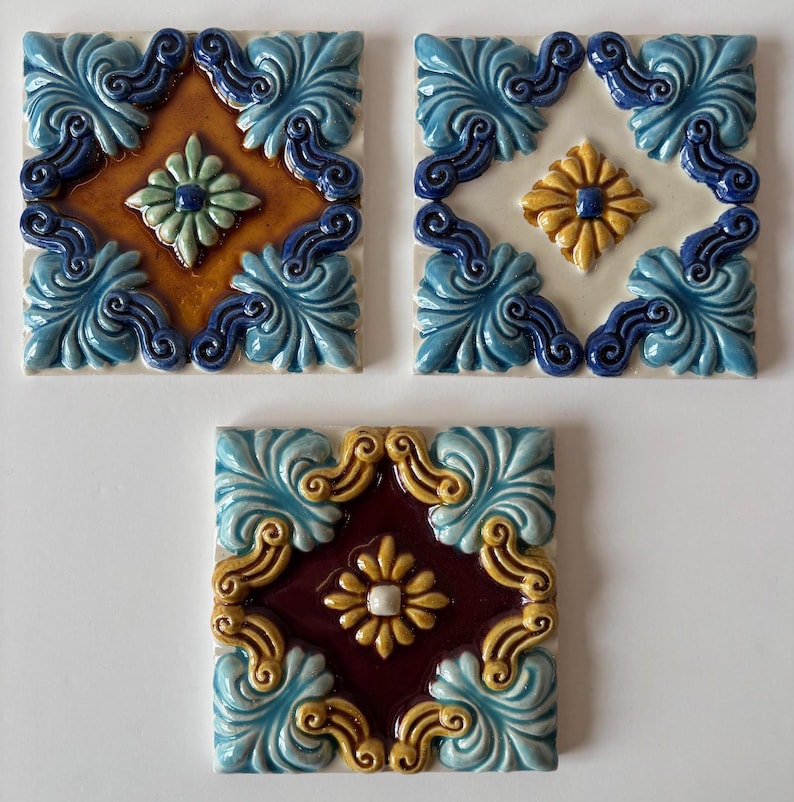 May include: Three square ceramic tiles with ornate designs. Each tile features a central floral motif in various colors, including yellow, green, and white, surrounded by a decorative border of blue and gold scrollwork. The tiles are likely for decorative use.