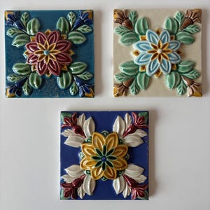 May include: Three square ceramic tiles with floral designs. One tile is teal with a maroon and yellow flower. Another is off-white with a blue and yellow flower. The third is blue with a yellow and white flower.