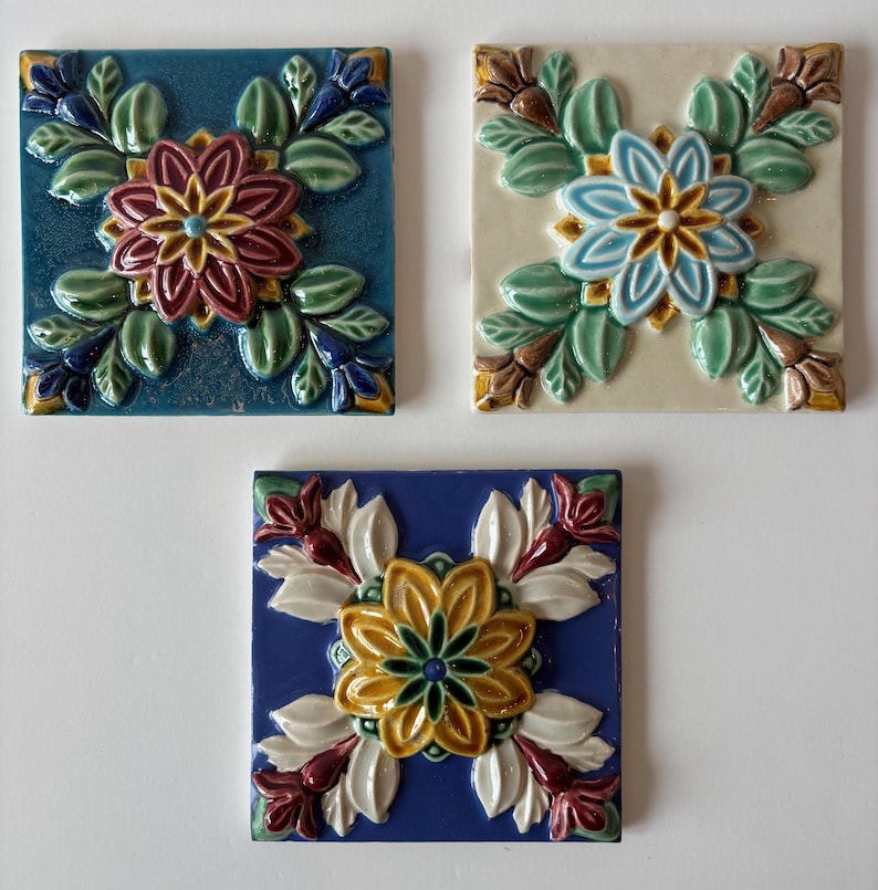 May include: Three square ceramic tiles with floral designs. One tile is teal with a maroon and yellow flower. Another is off-white with a blue and yellow flower. The third is blue with a yellow and white flower.