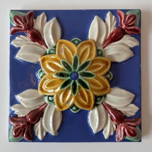 May include: Square ceramic tile with a raised floral design. The tile features a central yellow flower with green and blue accents, surrounded by white leaves and maroon flower buds, set against a blue background.