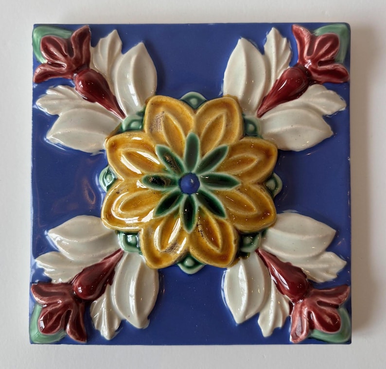 May include: Square ceramic tile with a raised floral design. The tile features a central yellow flower with green and blue accents, surrounded by white leaves and maroon flower buds, set against a blue background.