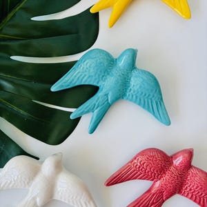 May include: Overhead shot of five ceramic swallows in various colors: yellow, turquoise, white, and red. The swallows are arranged on a white surface, with a large green leaf in the background. The swallows have detailed wing and tail feathers.