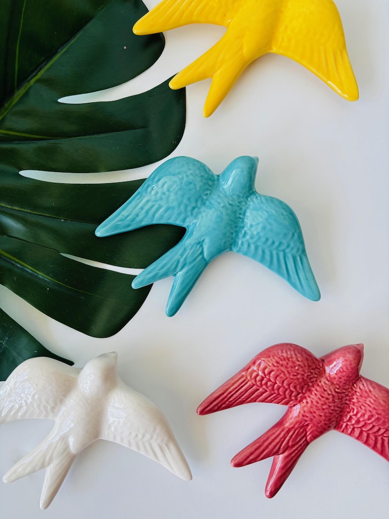 May include: Overhead shot of five ceramic swallows in various colors: yellow, turquoise, white, and red. The swallows are arranged on a white surface, with a large green leaf in the background. The swallows have detailed wing and tail feathers.