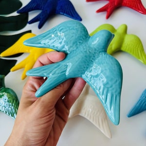 May include: A hand holding a turquoise ceramic bird figurine, with other colorful bird figurines in red, blue, yellow, green, and white. The birds have outstretched wings and are glossy.