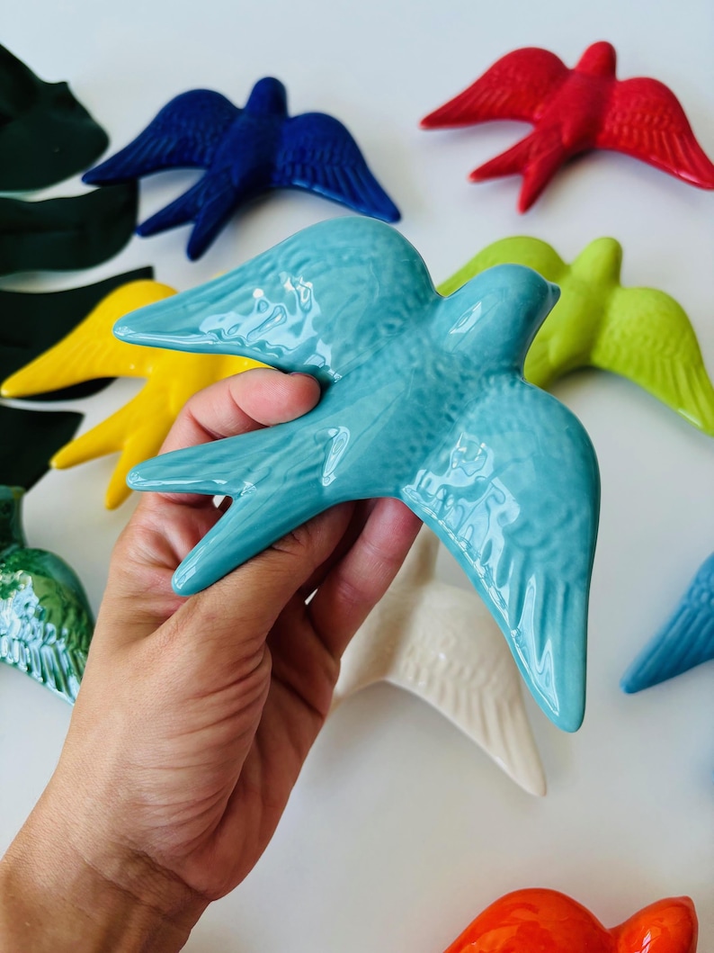 May include: A hand holding a turquoise ceramic bird figurine, with other colorful bird figurines in red, blue, yellow, green, and white. The birds have outstretched wings and are glossy.