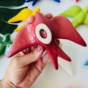 May include: A hand holds a glossy, red ceramic bird with a white circular detail. Other colorful ceramic birds in blue, yellow, green, and white are visible in the background. The birds have outstretched wings and a stylized design.