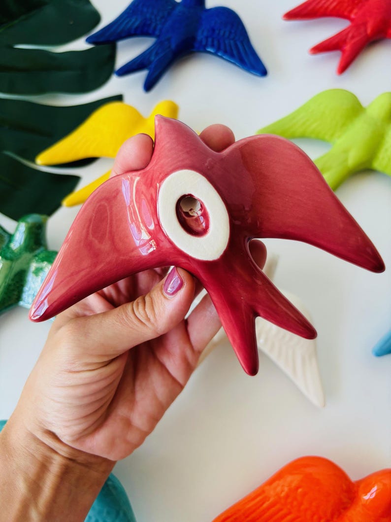 May include: A hand holds a glossy, red ceramic bird with a white circular detail. Other colorful ceramic birds in blue, yellow, green, and white are visible in the background. The birds have outstretched wings and a stylized design.