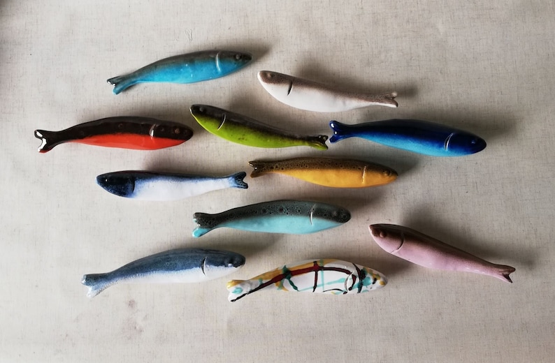 May include: A collection of twelve ceramic fish figurines in various colours, including blue, green, yellow, red, and pink. The fish are arranged in a semi-circle on a white background.