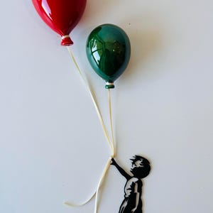 May include: A red and green balloon-themed art piece. The balloons are glossy and held by a black silhouette of a child. The balloons are red and green, with white ribbons. The child is holding the ribbons.