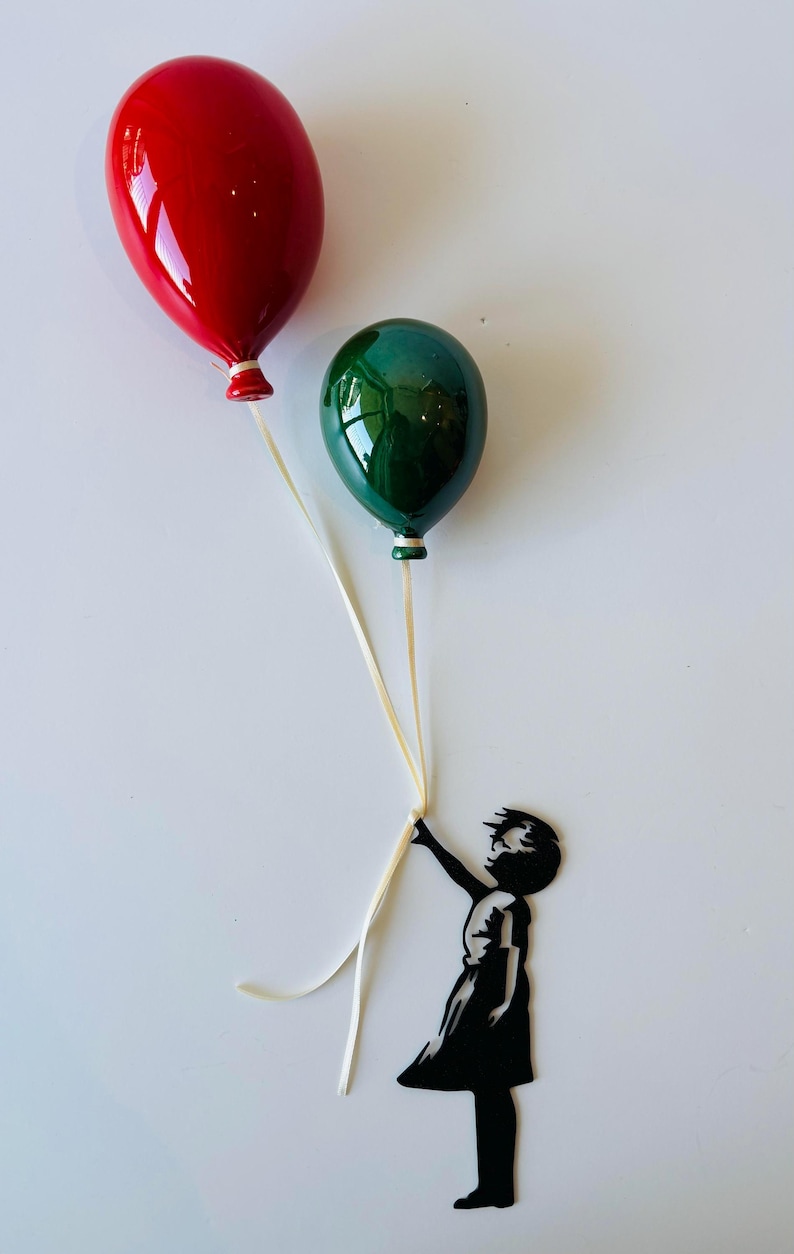 May include: A red and green balloon-themed art piece. The balloons are glossy and held by a black silhouette of a child. The balloons are red and green, with white ribbons. The child is holding the ribbons.