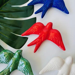 May include: A collection of ceramic swallow bird figurines in various colors: blue, red, green, and white. The birds have outstretched wings and are arranged on a white surface with a large green leaf in the background. These decorative birds are suitable for home decor.