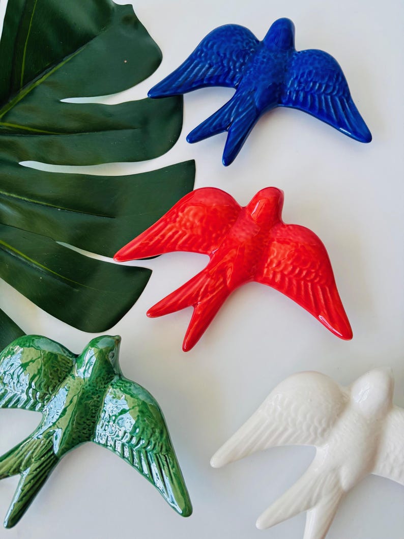 May include: A collection of ceramic swallow bird figurines in various colors: blue, red, green, and white. The birds have outstretched wings and are arranged on a white surface with a large green leaf in the background. These decorative birds are suitable for home decor.