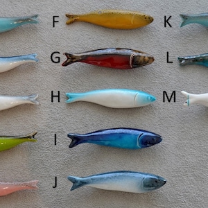 May include: Twelve ceramic fish figurines in various colours and patterns, including blue, green, red, yellow, and white. The fish are arranged in four rows of three, with letters A through M labelling each fish.