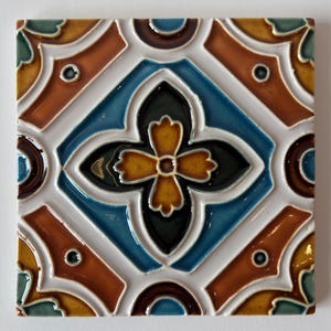 May include: Square ceramic tile with a repeating geometric pattern. The design features a central blue diamond with a yellow and black floral motif. The tile has brown, yellow, and teal accents, outlined in white.