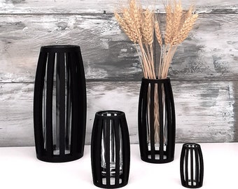 May include: Four black vases with a vertical slat design. The largest vase is on the left and the smallest is on the right. The middle vase holds a bouquet of dried wheat.