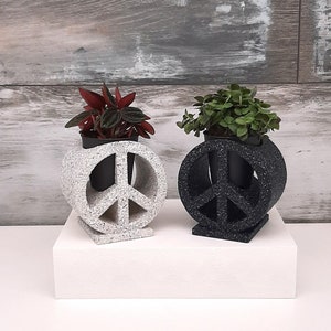May include: Two peace sign shaped planters, one white and one black, with small plants inside. The white planter has a green plant and the black planter has a red plant.