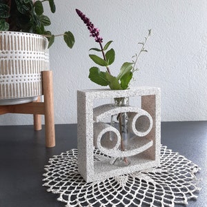 Puede incluir: Un jarrón de cerámica blanca con forma de signo astrológico Cáncer. El jarrón tiene un tubo de vidrio transparente en el interior para sostener flores. Una rama de flores moradas y hojas verdes está en el jarrón.