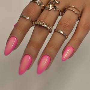Könnte beinhalten: Eine Hand mit mehreren goldenen Ringen in verschiedenen Designs, darunter ein wellenförmiges Band, ein blütenförmiger Ring und ein Doppelbandring. Die Nägel sind lang, oval und mit einem rosa-weißen Farbverlauf lackiert.