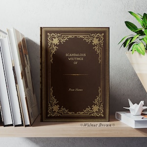 May include: A brown book with gold accents and the title "Scandalous Writings of Fran Hants" on the cover. The book is on a wooden shelf with other books and a hanging plant.