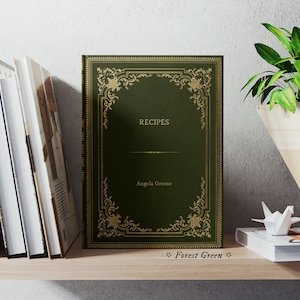 May include: A dark green hardcover book with gold accents and the title "RECIPES" on the front cover. The author's name, "Angela Greene", is printed below the title. The book is on a wooden shelf with other books and a hanging plant.