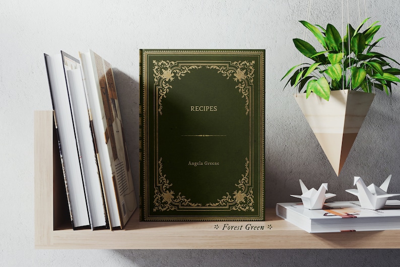 May include: A dark green hardcover book with gold accents and the title "RECIPES" on the front cover. The author's name, "Angela Greene", is printed below the title. The book is on a wooden shelf with other books and a hanging plant.
