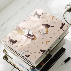 May include: A stack of notebooks with a pink cover featuring a repeating pattern of black cats, moons, and stars. The cover has a vintage aesthetic.