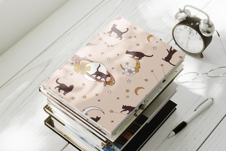 May include: A stack of notebooks with a pink cover featuring a repeating pattern of black cats, moons, and stars. The cover has a vintage aesthetic.
