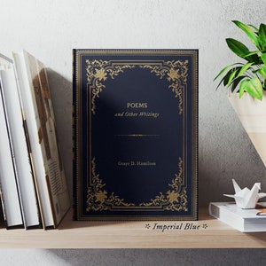 May include: A navy blue hardcover book with gold lettering and a decorative gold frame. The title is "POEMS and Other Writings" by "Graye D. Hamilton". The book is on a wooden shelf with other books and a hanging plant.