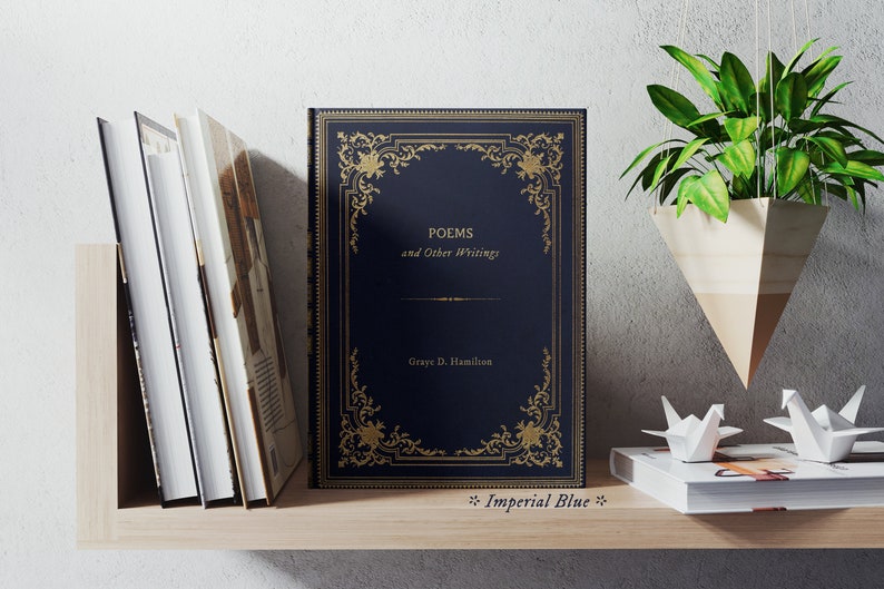 May include: A navy blue hardcover book with gold lettering and a decorative gold frame. The title is "POEMS and Other Writings" by "Graye D. Hamilton". The book is on a wooden shelf with other books and a hanging plant.