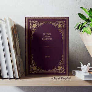 May include: A purple book with gold lettering and a decorative gold frame on the cover. The title of the book is "Letters to My Daughter" and the author is "Darla". The book is on a wooden shelf with other books and decorative items.