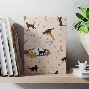 May include: A pink notebook with a repeating pattern of black cats, stars, moons, and flowers. The notebook is on a shelf with other books and a hanging plant.