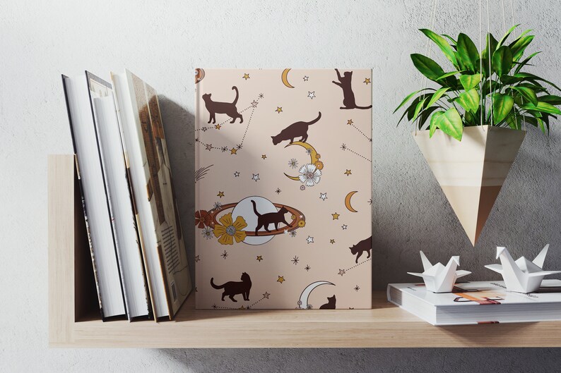 May include: A pink notebook with a repeating pattern of black cats, stars, moons, and flowers. The notebook is on a shelf with other books and a hanging plant.